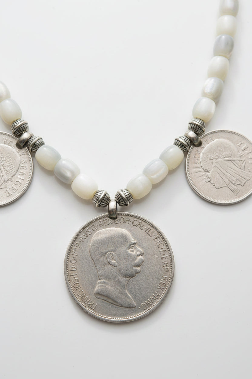 A close-up of a beaded necklace with white shell-like beads and silver-toned spacer beads, featuring a coin pendant and another coin laid on a dark green textured fabric background.
