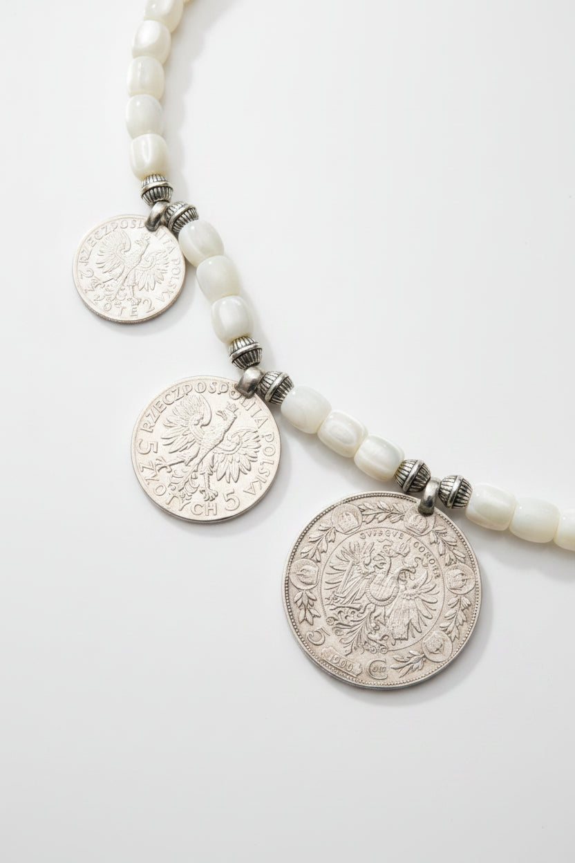 A close-up of a beaded necklace featuring three silver-toned coin pendants and white oval beads, laid flat on a textured dark green fabric background.