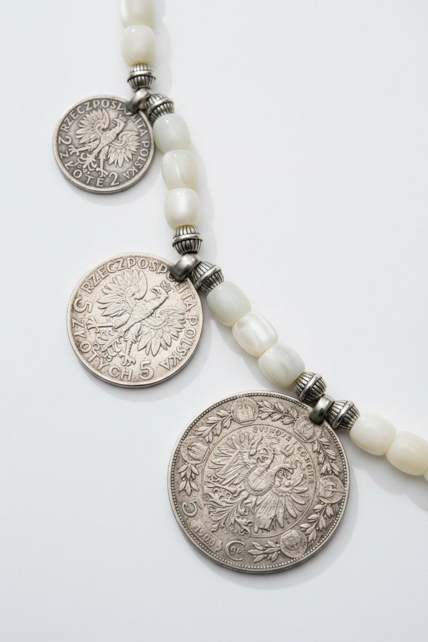 A close-up of a beaded necklace featuring three silver-toned coin pendants and white oval beads, laid flat on a textured dark green fabric background.