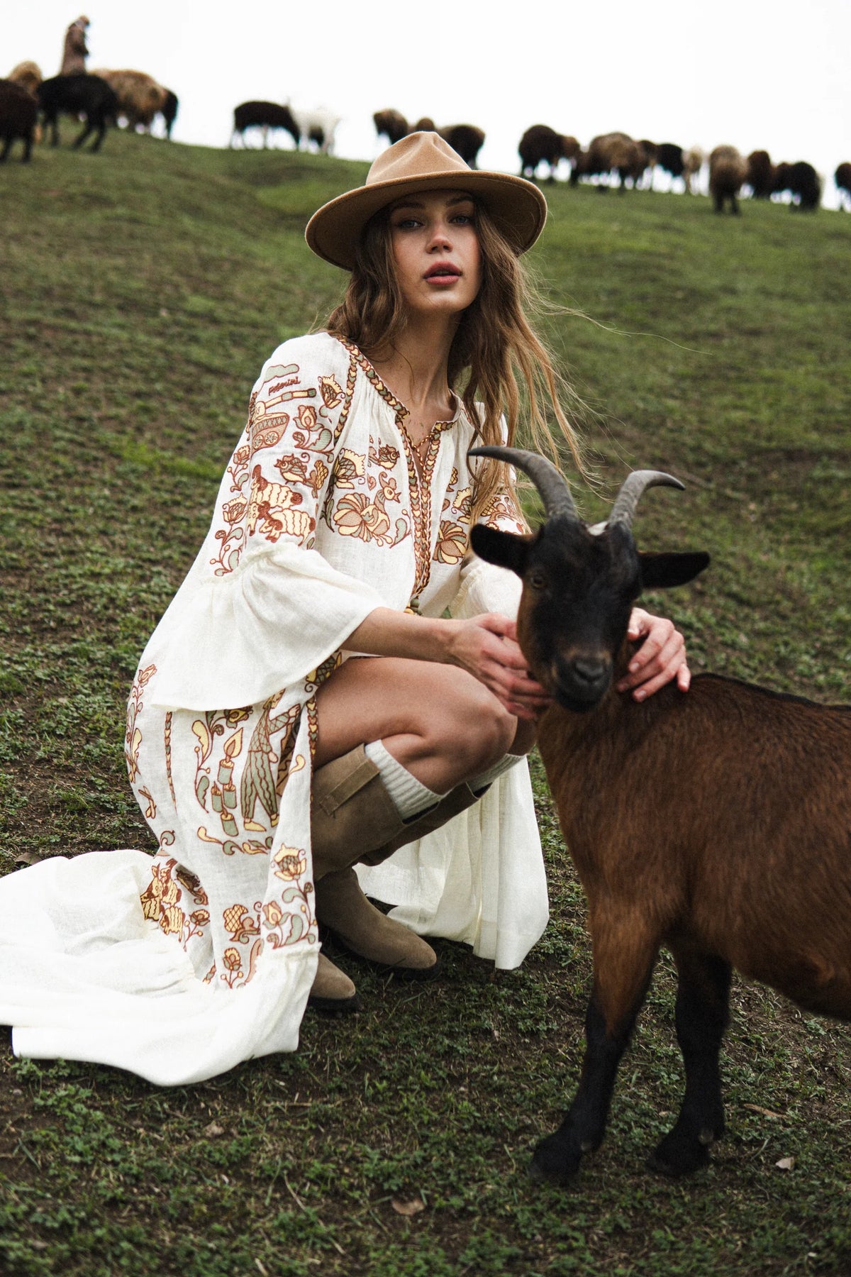 Woman in a white embroidered dress and hat with a goat on a grassy hill with grazing animals in the background