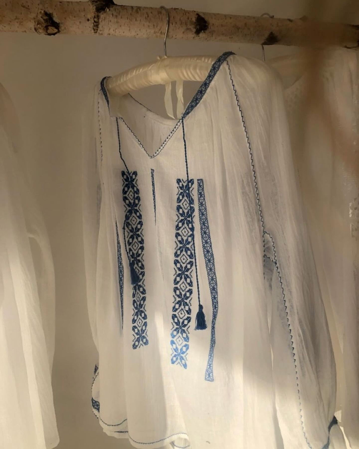 White garment with blue embroidery hanging on a wooden stick against a white background