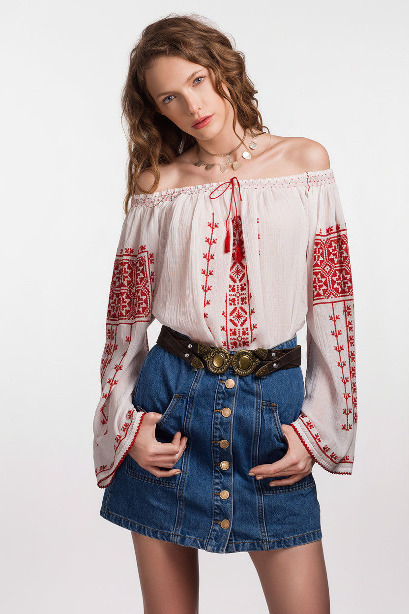Off-shoulder hand-embroidered Romanian blouse ia with traditional Transylvanian red cross-stitch motifs on white cotton gauze, worn on a misty beach.