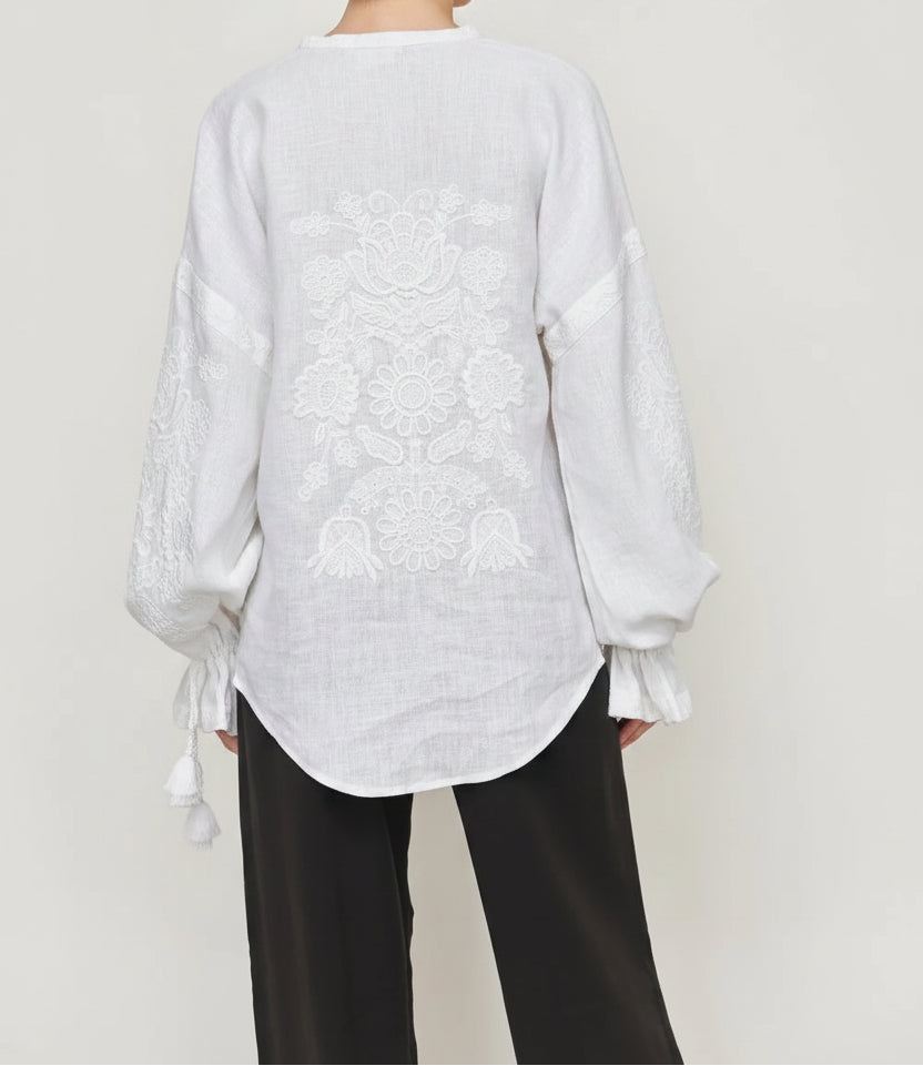 Woman white long-sleeve linen blouse with intricate floral embroidery and tie details at the cuffs, paired with black pants. Blouse Roumaine Shop