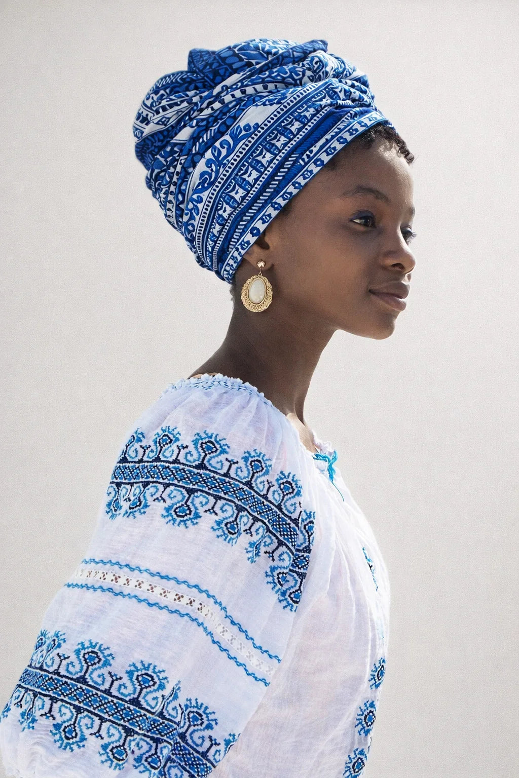 Woman wearing a white outfit with blue embroidery and a matching headscarf against a plain background