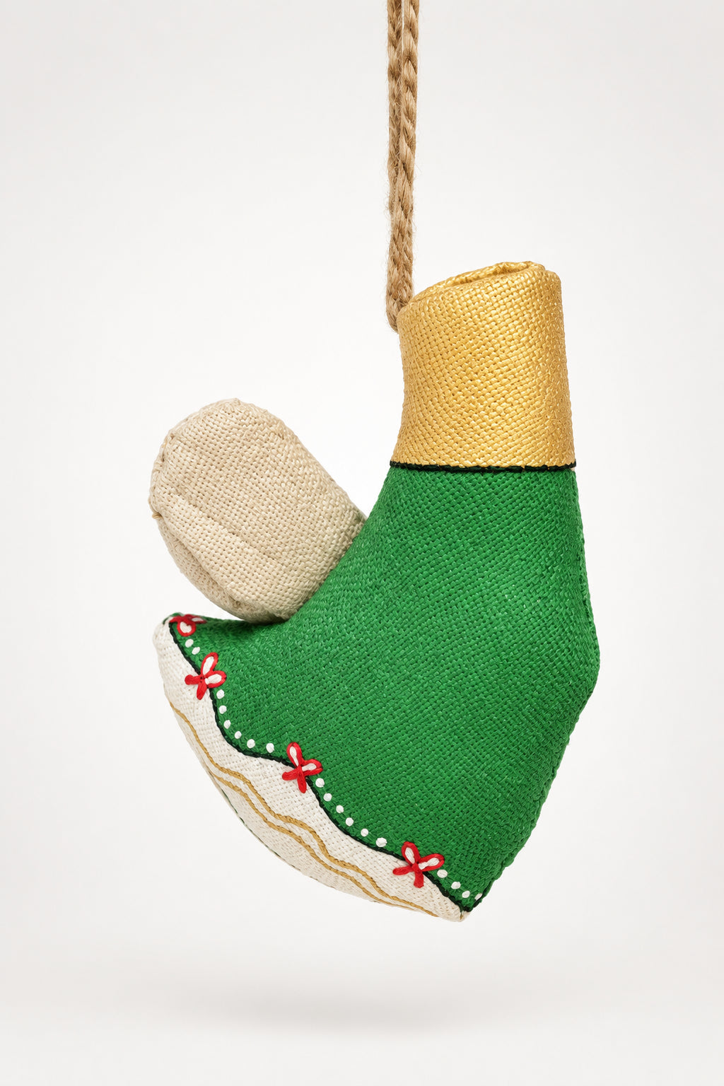 A handmade Christmas ornament shaped like an elf's boot hangs from a pine tree branch. The boot is made from green burlap fabric with a tan burlap cuff, a beige fabric wing, and small red embroidered details along the sole.