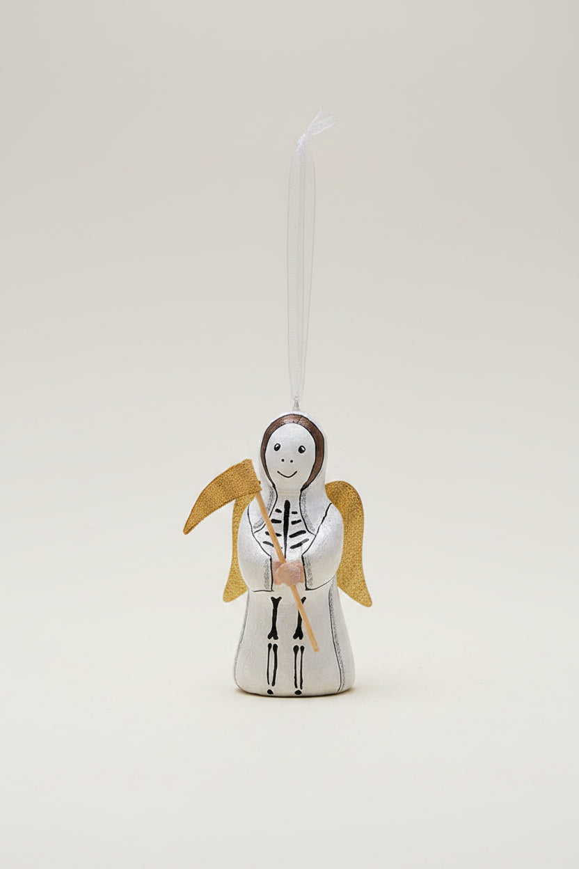 A hand-painted wooden Christmas angel ornament with a white robe, black skeletal details, and a gold halo, hanging from a clear ribbon under pine branches against a dark green background and pale yellow surface.
