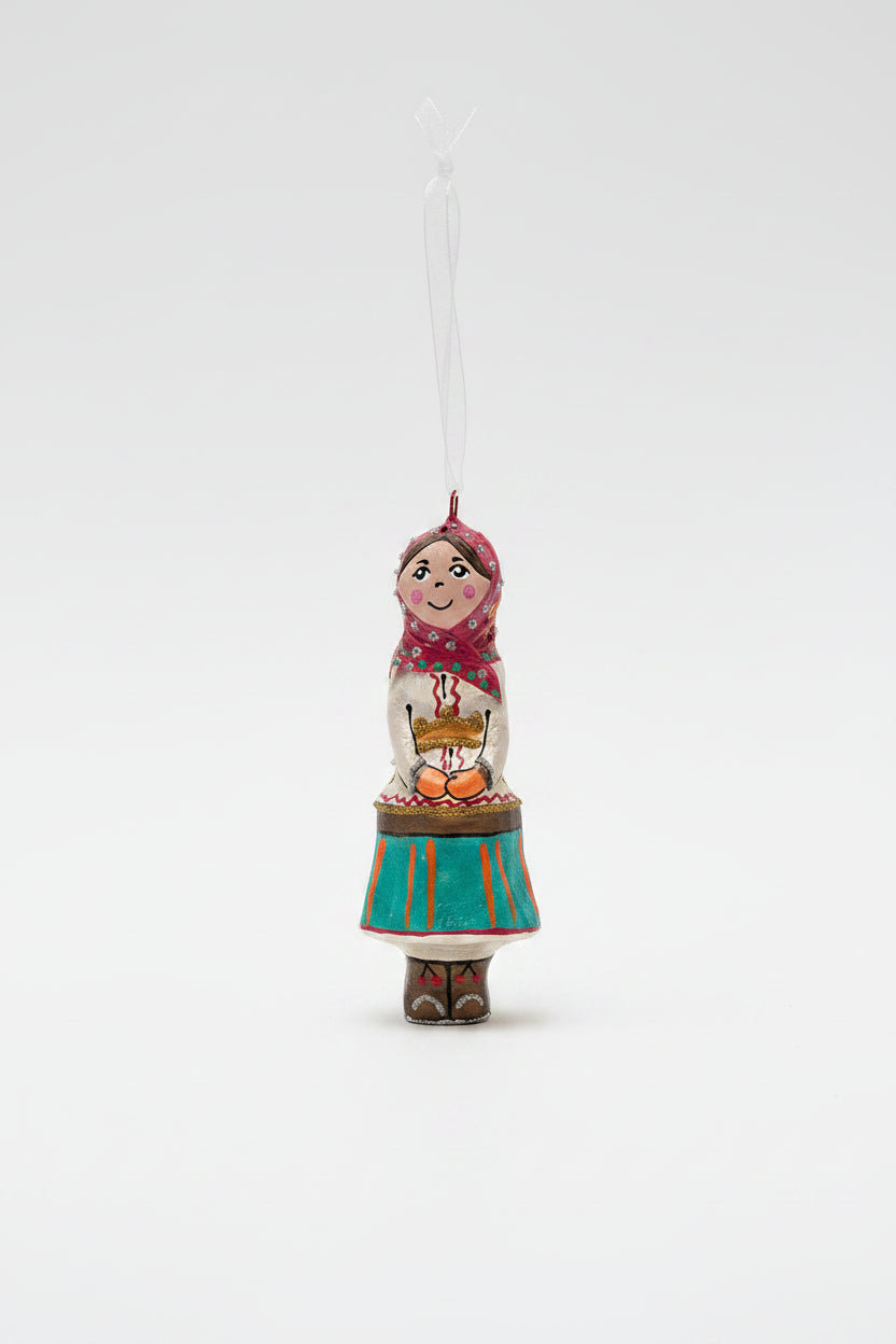 A hand-painted wooden Christmas ornament shaped like a woman in traditional Russian attire, hanging from a pine branch against a dark green background and cream surface.