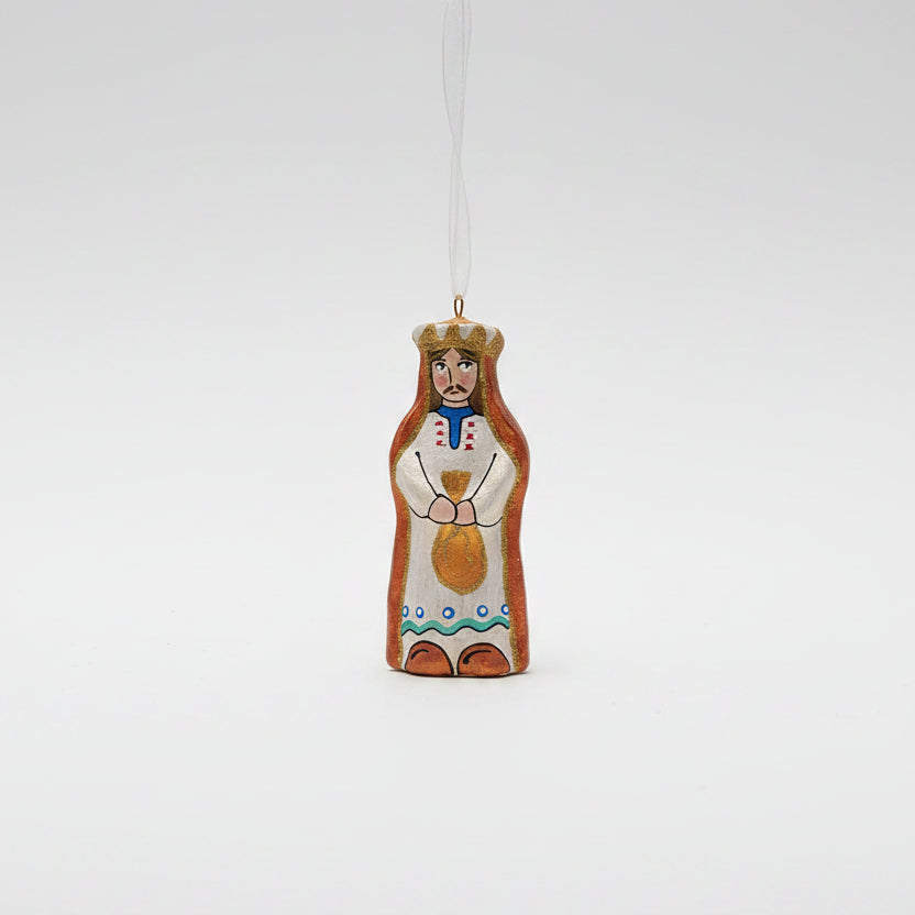 A hand-painted wooden Christmas ornament shaped like a crowned king in traditional folk clothing, hanging from a pine branch against a dark green background and cream surface.