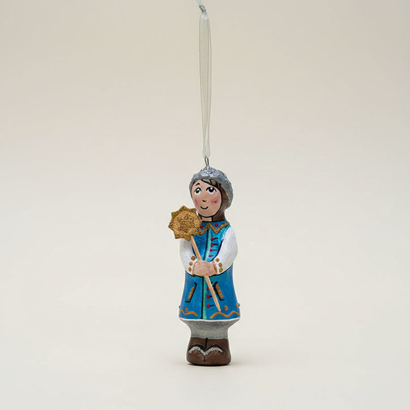 A hand-painted wooden Christmas ornament shaped like a traditional Slavic figure in blue and white clothing holding a star, hanging from a pine branch against a dark green background and beige surface.