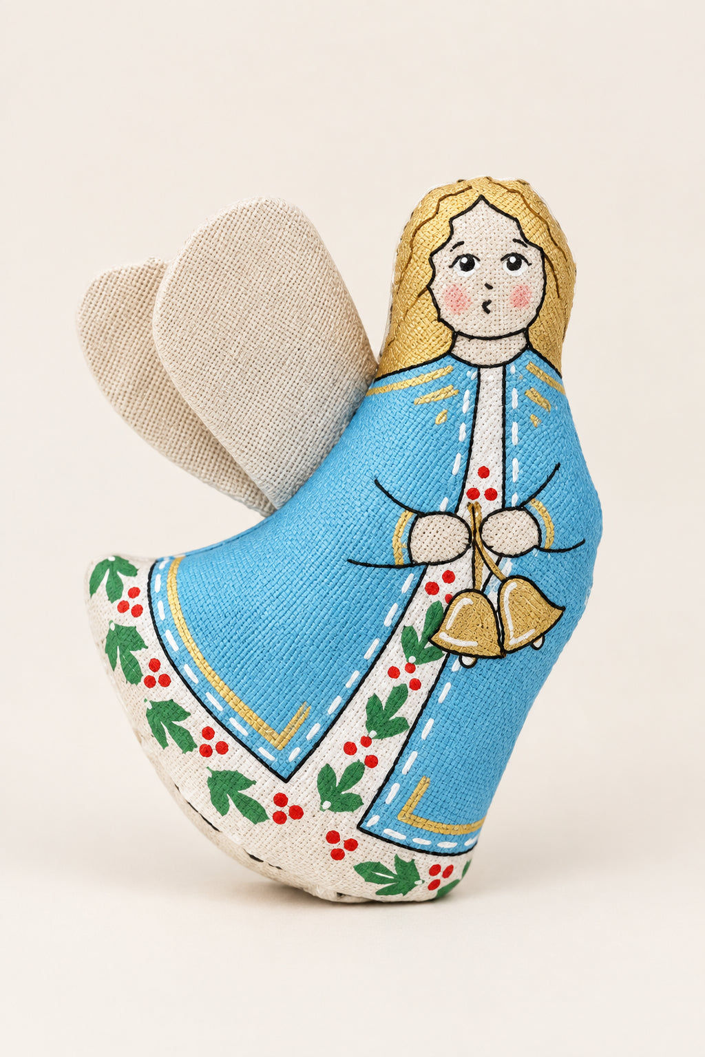 A handmade fabric angel Christmas ornament with a light blue dress, gold hair, and green holly accents, nestled in white snow with pine branches and silver ornaments in the background.