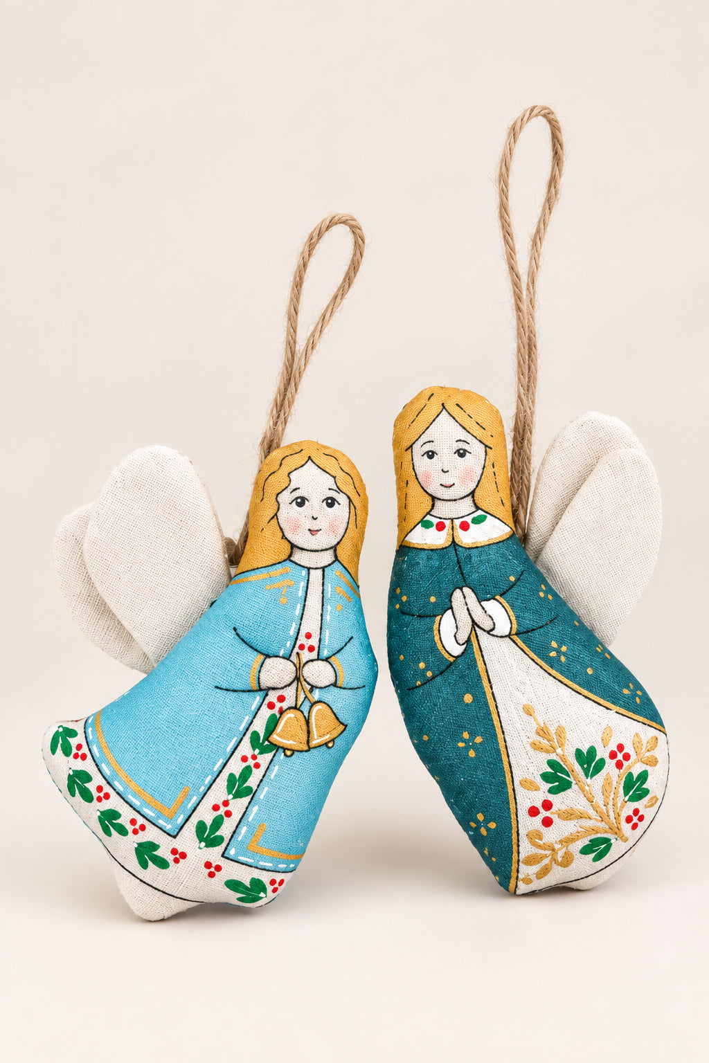 Two handmade fabric angel ornaments with white wings, hanging from twine, displayed on a brown felt background. One angel wears a light blue dress with green and red floral patterns, and the other wears a teal dress with gold stars and a red collar.