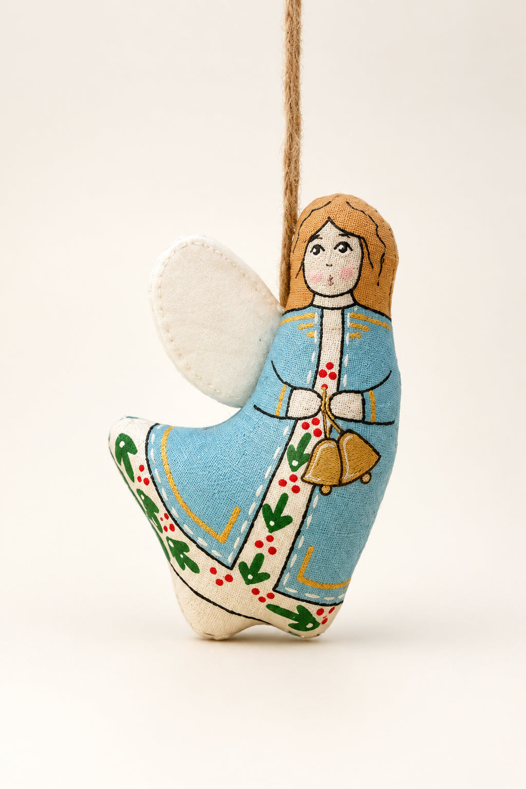 A small hand-painted wooden Christmas angel ornament hangs from a pine branch against a dark green background and pale yellow surface.