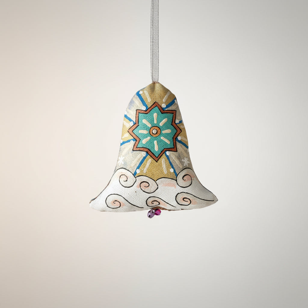 A decorative fabric Christmas ornament shaped like a bell hangs from a pine tree branch. The ornament has a beige linen-like background with a central teal and orange geometric star motif, blue rays, and white cloud-like swirls along the bottom edge. Blouse Roumaine Shop