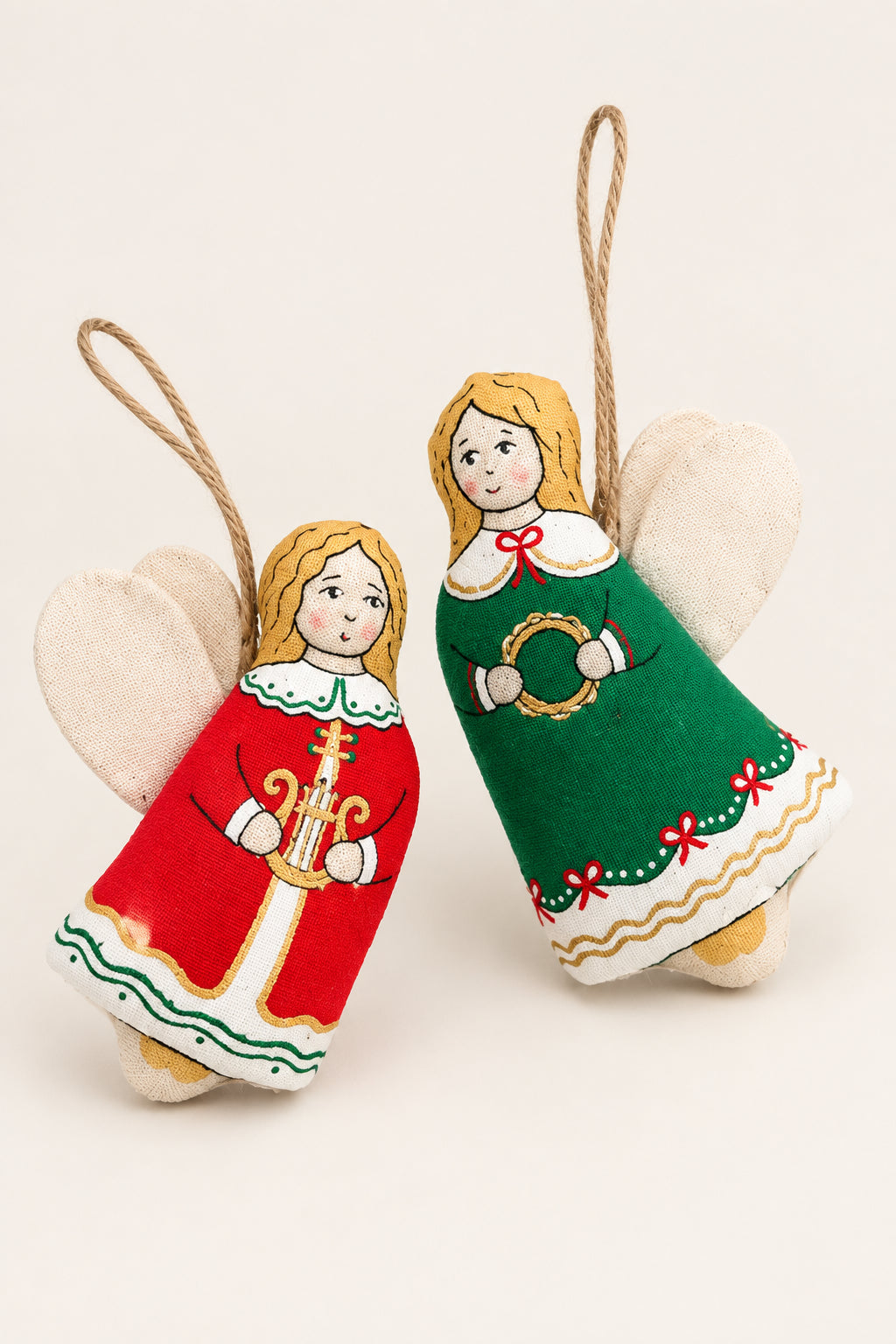Two handmade fabric Christmas angel ornaments with white wings, hanging from twine, displayed on a brown textured background. One angel wears a red dress with gold and white details, and the other wears a green dress with red and gold details.