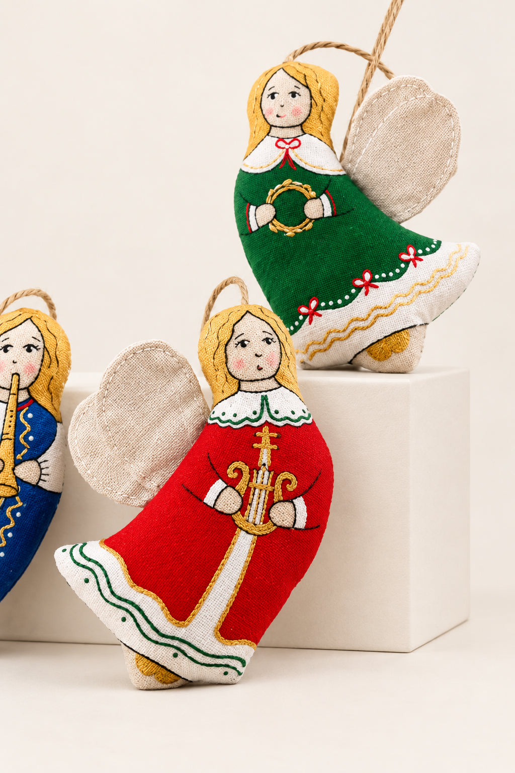 Three embroidered Christmas angel ornaments with fabric wings, displayed in front of patterned brown gift boxes on a rustic surface.
