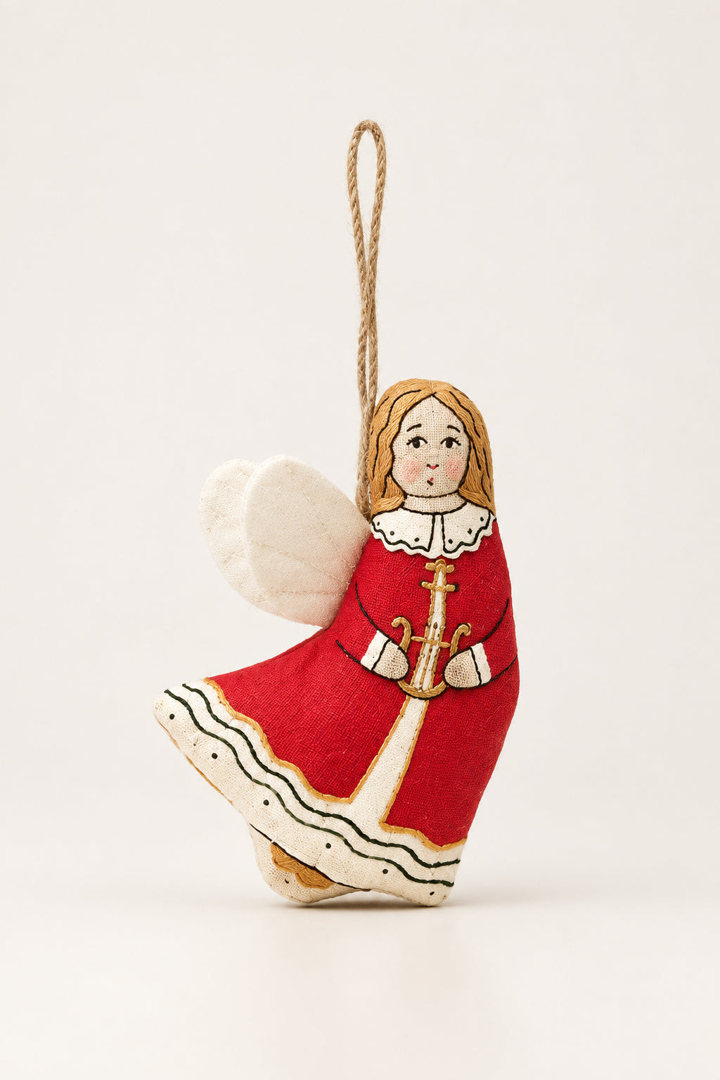 A handcrafted wooden Christmas angel ornament wearing a red dress with white and gold details, hanging from a pine branch against a dark green background and pale yellow surface.