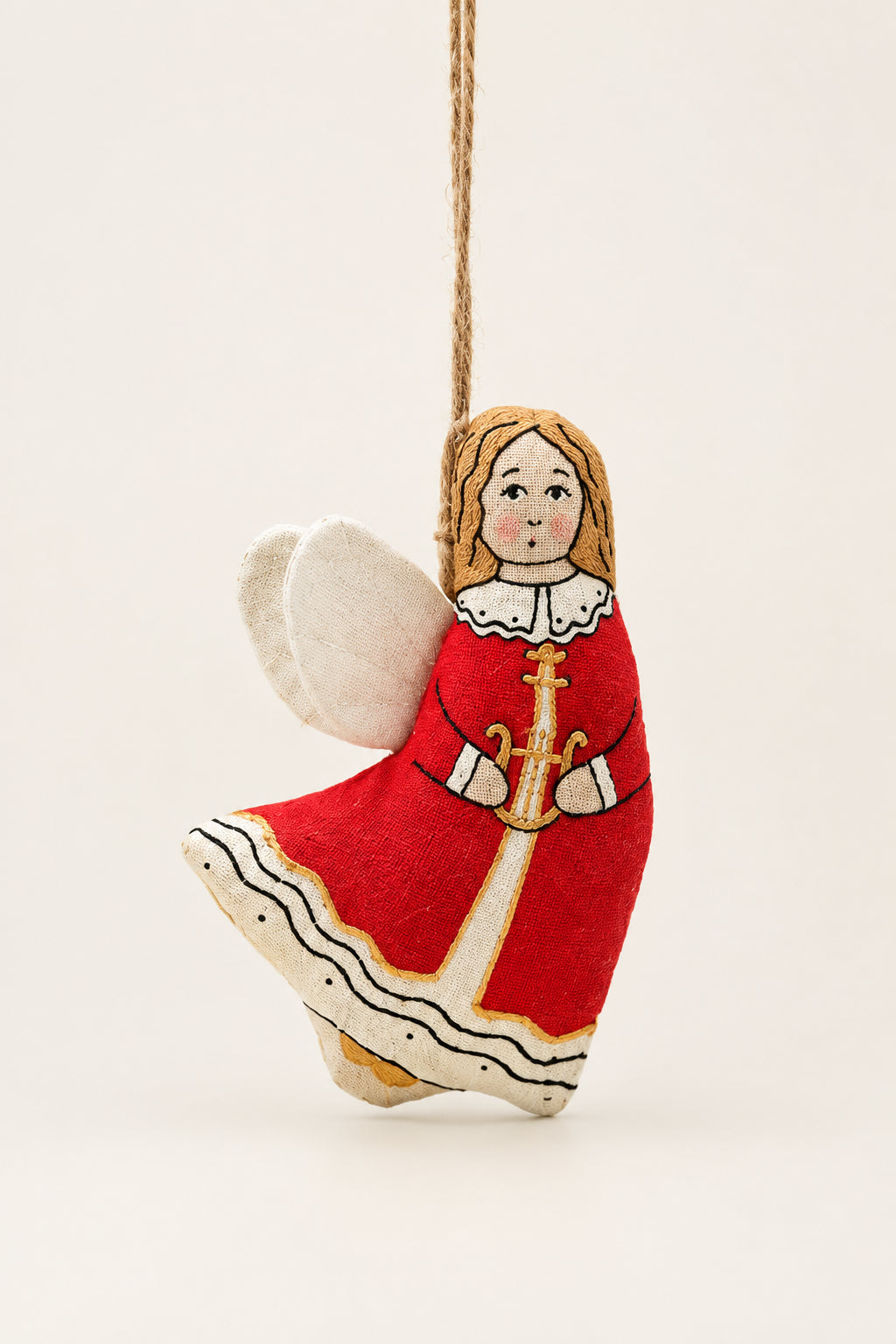A hand-painted wooden Christmas angel ornament wearing a red dress with white and gold details, hanging from a pine branch against a dark green background and a pale yellow surface.