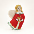 A handmade embroidered angel Christmas ornament in a red dress with gold and white details, standing in white snow with rustic holiday decorations blurred in the background.