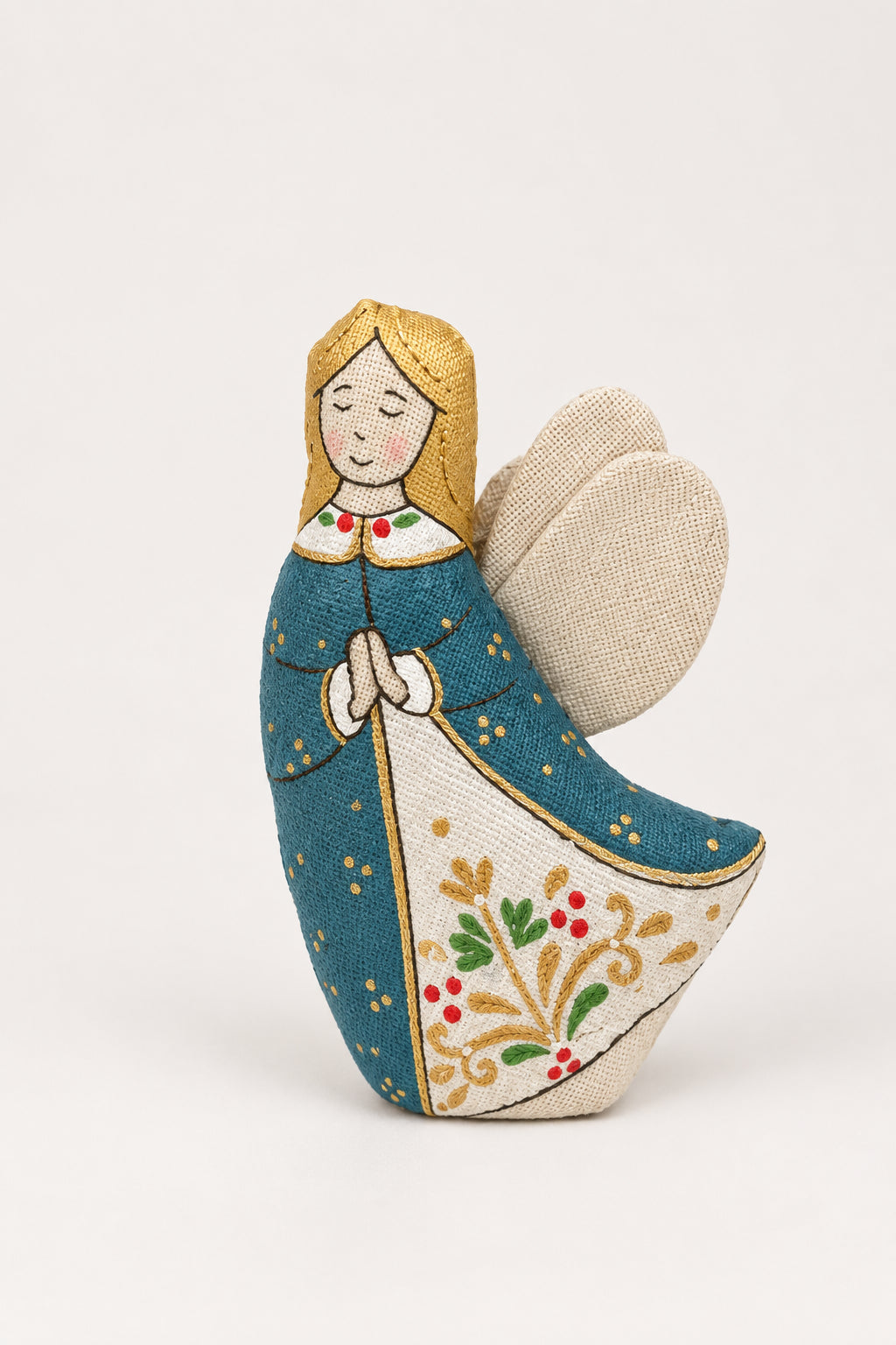 A handmade fabric Christmas angel ornament standing in white snow, with pine branches, silver ornaments, and a blue textured background.