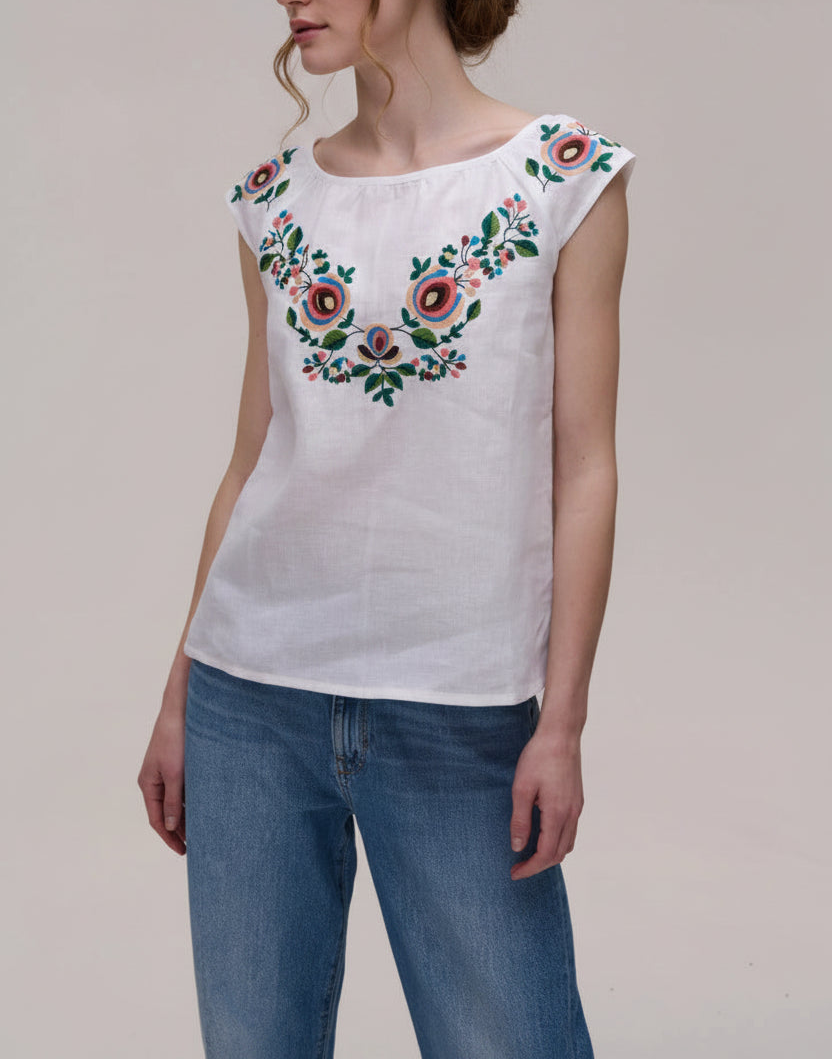A woman wearing a white sleeveless top with colorful floral embroidery across the chest and blue jeans, photographed against a plain light beige studio background.