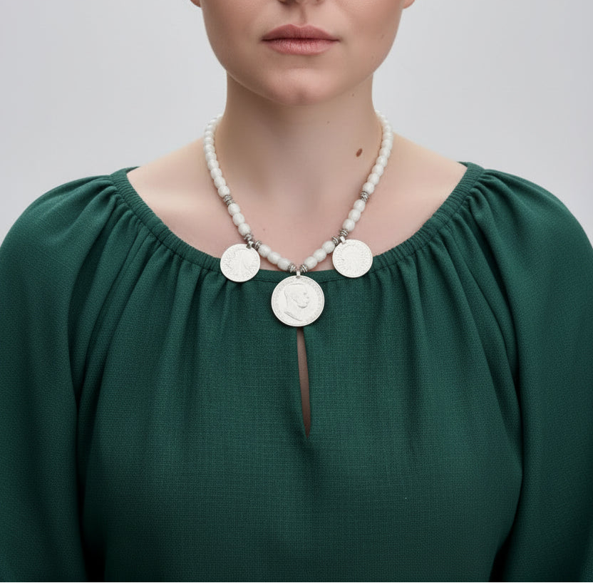 A close-up of a model wearing a necklace made of white pearl-like beads and silver coin charms, paired with a dark green off-the-shoulder top.
